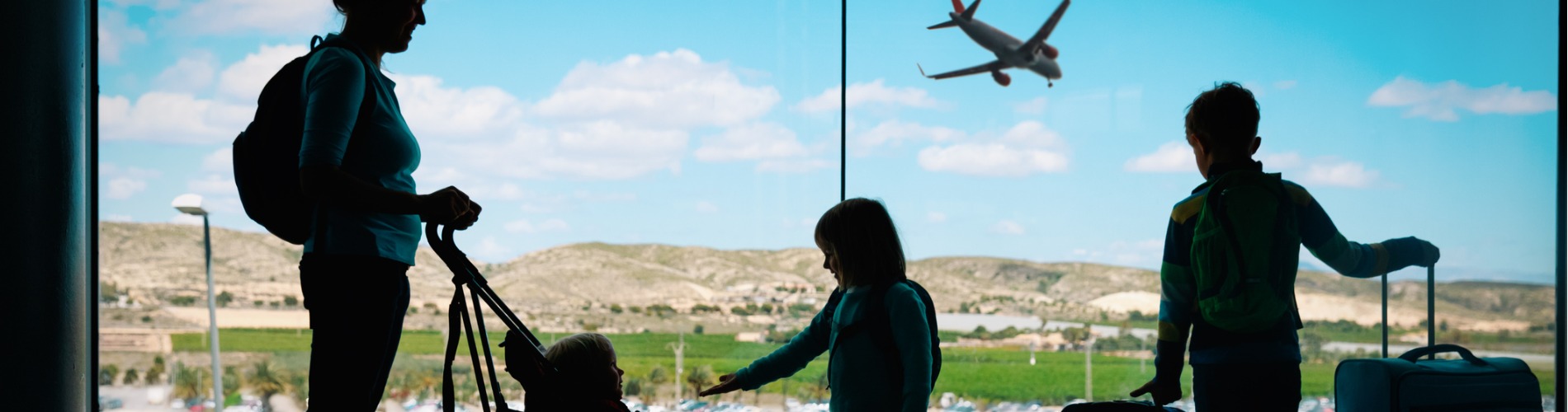 mother-with-kids-and-luggage-looking-at-planes-in-airport 1900 x 500